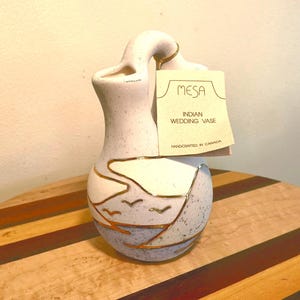 Peut inclure: Un vase de mariage indien en céramique avec une forme unique et une anse incurvée. Le vase est principalement blanc avec des accents bleus et dorés, avec un motif de paysage avec des oiseaux. L'étiquette indique "MESA INDIAN WEDDING VASE HANDCRAFTED IN CANADA."