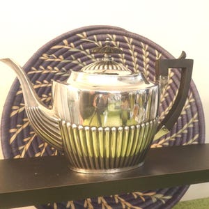 Puede incluir: Una tetera plateada con un asa y un pomo de tapa de color marrón oscuro. La tetera tiene un diseño acanalado en la mitad inferior y un pico curvo. Se encuentra sobre una superficie oscura con un fondo tejido azul y blanco.