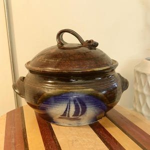May include: A ceramic lidded casserole dish with a sailboat design. The dish is primarily brown with blue accents and a white sailboat image. It has a curved handle on the lid and two side handles.