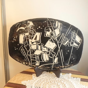 Peut inclure: Un plat en céramique noir avec un motif abstrait blanc. Le motif présente des formes géométriques et des lignes, créant une esthétique moderne et artistique. Le plat est présenté sur un support en bois, mettant en valeur son caractère décoratif.