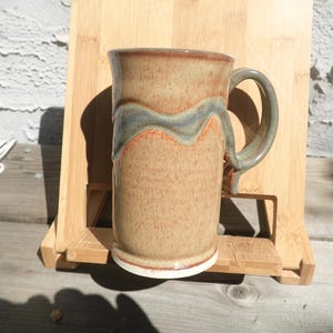 May include: A handmade ceramic mug with a speckled tan body, a wavy blue and orange design, and a curved handle. The mug has a white base and a brown rim. The mug is displayed on a wooden stand.