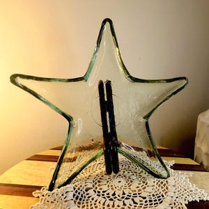 May include: A clear glass star-shaped dish with a slightly green tint. The dish is sitting on a white crocheted doily on a wooden surface.