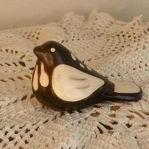 Puede incluir: Una figurita de pájaro de cerámica pintada en blanco y negro. El pájaro tiene un cuerpo redondeado y un pico largo y puntiagudo. El pájaro está sentado sobre una superficie de crochet blanco.