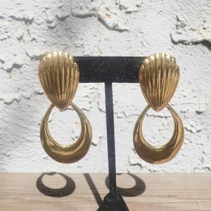 May include: A pair of gold-coloured earrings. Each earring has a textured, fan-shaped top and a large, teardrop-shaped dangle. The earrings are displayed on a black stand against a textured white and grey background.