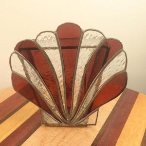 May include: A stained glass seashell sculpture with alternating red and clear glass panels. The decorative piece is set on a wooden surface with a striped pattern. The sculpture is approximately 15 cm tall.