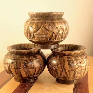 May include: Three hand-carved wooden pots with a dark brown finish. Each pot has a rounded shape and is decorated with carved relief patterns. The pots are stacked, creating a visually appealing arrangement.