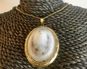 Op de afbeelding: Een ovaal gouden medaillon met een witte en grijze schelp cameo hanger aan een gouden ketting.