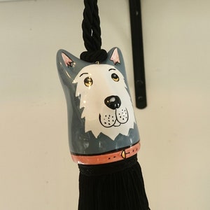 Könnte beinhalten: Ein grauer Keramik-Tasselauftrag in Form eines Hundekopfes mit einer schwarzen Schnur und einem schwarzen Quasten. Der Hund hat ein weißes Gesicht mit schwarzen Augen und einer schwarzen Nase.