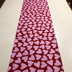 May include: A rectangular fabric table runner featuring a repeating pattern of light pink hearts set against a deep red background. The runner has a stitched border along all four edges, creating a clean, finished look for this textile home decor item.
