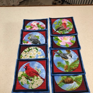 May include: Set of eight square fabric coasters with a blue border and red center. Each coaster features a different bird and floral design.