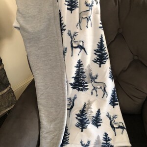 May include: A soft, white fleece blanket with a pattern of dark blue trees and deer. One side of the blanket is gray.