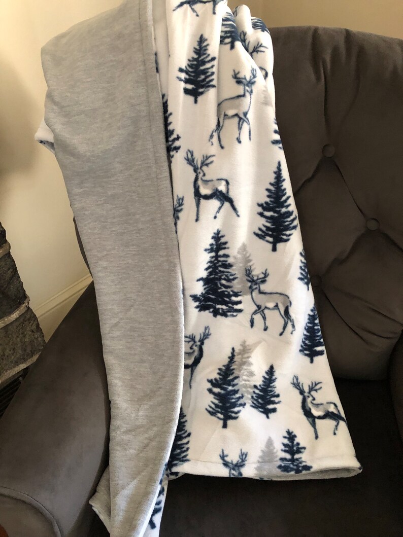 May include: A soft, white fleece blanket with a pattern of dark blue trees and deer. One side of the blanket is gray.