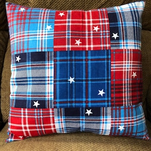 May include: Square pillow with a patchwork design featuring red, white, and blue plaid patterns. White star accents are scattered across the pillow's surface, creating a patriotic theme. The pillow is resting on a brown surface.