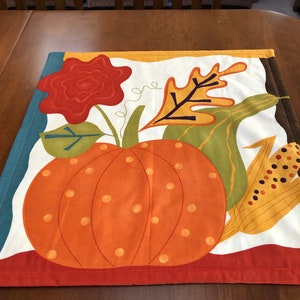 May include: A white fabric table runner with a colorful autumn-themed design. The design features a large orange pumpkin with brown stems, a red flower, green leaves, and a yellow ear of corn. The runner has a red, orange, and yellow border.