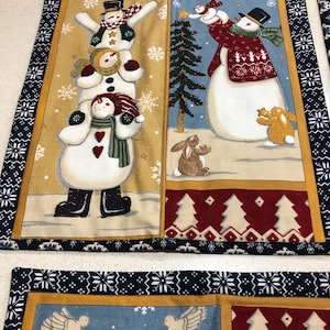 May include: Two sets of Christmas-themed placemats with a blue, red, and gold color scheme. The first set features a snowman family with a Christmas tree and a bunny. The second set features a snowman with two birds and a Christmas tree.