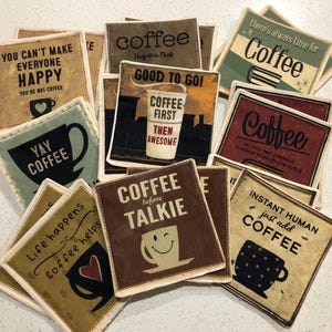 May include: A collection of square coffee coasters with various designs and text. The coasters feature phrases like "Coffee First Then Awesome" and "Instant Human Just Add Coffee." Colors include brown, beige, green, and black. The coasters have a stitched border.