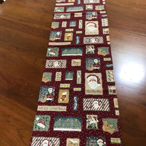 May include: A rectangular fabric table runner featuring a deep red background with a white speckled pattern. The design includes various rectangular patches depicting Santa Claus, Christmas trees, teddy bears, stockings, and the text Merry Christmas, Joy, and Ho Ho Ho.