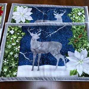 May include: Two placemats with a winter theme. The placemats feature a blue background with white snowflakes, a white poinsettia, and a gray silhouette of a deer. The placemats have a green border with a white snowflake pattern.