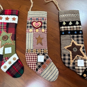 May include: Three fabric Christmas stockings with hanging loops. Stocking 1 features red, green, and white plaid with a green star and buttons. Stocking 2 has a heart, star, and buttons on plaid fabric. Stocking 3 is brown plaid with a star and buttons.