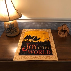 May include: A beige and brown Christmas-themed wall hanging with a silhouette of three camels and a star. The text "Joy to the World" is written in red and gold lettering.