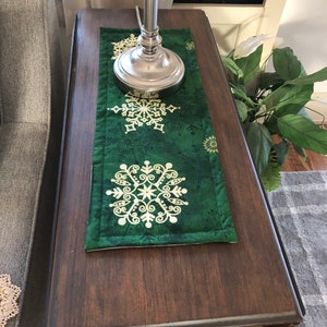 May include: A green table runner with a gold snowflake design. The runner is on a dark wood table with a silver lamp on top.