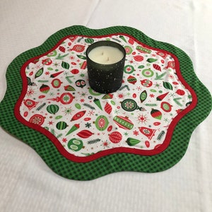 May include: A scalloped fabric mat featuring a white background with a pattern of red and green Christmas ornaments, pine needles, and snowflakes. The mat has a red inner border and a green and black gingham outer edge, topped with a dark candle.