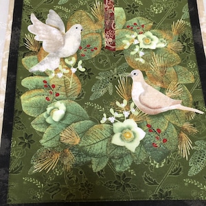 May include: A green fabric wall hanging with a wreath design featuring two white doves, white flowers, and red berries. The wreath is surrounded by a dark green border.