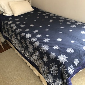 May include: A navy blue twin-sized bedspread with a white snowflake pattern. The bedspread has a white lace trim and is quilted.