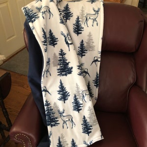 May include: A white fleece throw blanket with a navy blue and gray pattern of trees and deer. The blanket is folded and draped over a brown leather armchair.