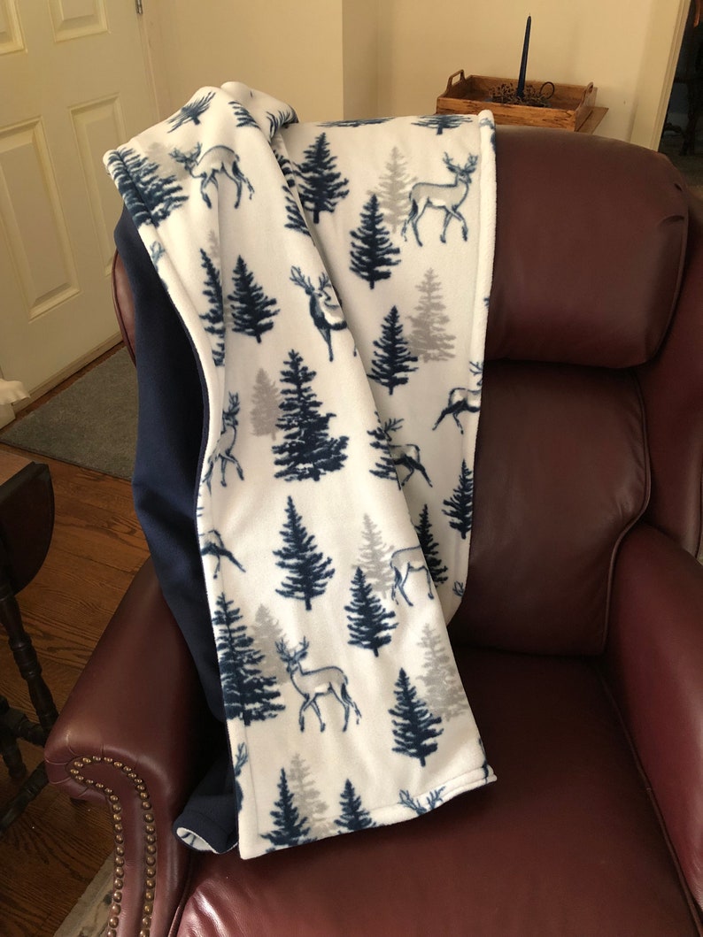 May include: A white fleece throw blanket with a navy blue and gray pattern of trees and deer. The blanket is folded and draped over a brown leather armchair.