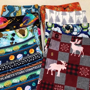 May include: A collection of folded fabric pieces with various patterns. Designs include planets, dinosaurs, basketballs, Christmas trees, soccer balls, and reindeer. The fabrics feature a mix of colors, including blue, orange, and red, with some text visible.