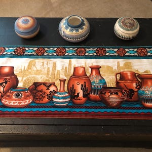 May include: A rectangular table runner featuring a desert landscape print with various terracotta-colored pottery vessels, cacti, and geometric borders in turquoise, orange, and black. Three small, round, decorative lidded jars sit above it.