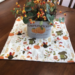 May include: A white table runner with a fall-themed design featuring turkeys, squirrels, acorns, leaves, and pumpkins. The runner has a brown border and is decorated with a metal container with the text "Pumpkin Pickin' Local Farm Fresh".