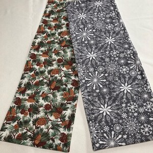 May include: Two fabric table runners with a winter holiday theme. One runner features a white background with a green pine branch and brown pine cone pattern. The other runner features a gray background with a white snowflake pattern.
