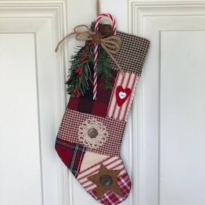 May include: A handmade Christmas stocking with a patchwork design in red, white, and brown fabrics. It features a heart, star, and lace embellishments, and is decorated with a candy cane, greenery, and a small bell. The stocking hangs from a jute twine loop.