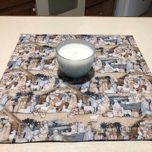 May include: A light blue glass candle holder with a white candle sits on a rectangular fabric placemat. The placemat features a detailed print depicting a nativity scene with figures, animals, and buildings in muted tones.
