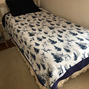 May include: A white twin-sized fleece blanket with a navy blue and gray pattern of stylized trees and deer. The blanket has a navy blue and white crocheted trim.