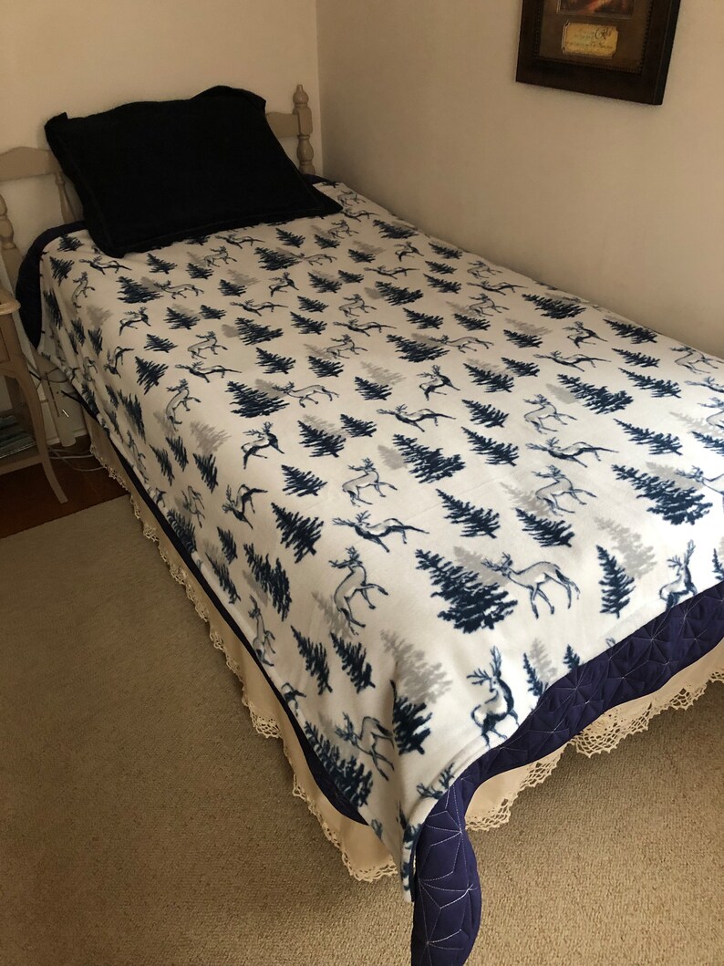 May include: A white twin-sized fleece blanket with a navy blue and gray pattern of stylized trees and deer. The blanket has a navy blue and white crocheted trim.