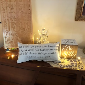 May include: A wooden sign with religious text, a small pillow with a Bible verse, a lit wooden block with "Peace & Love", and a glass angel figurine. Decorative lights are strung across the items.