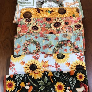 May include: A stack of colorful fabric pieces with autumn-themed patterns. Designs include sunflowers, fall leaves, wreaths, and mushrooms. The fabrics feature a variety of colors, including yellow, orange, green, and blue. Some fabrics have text printed on them.