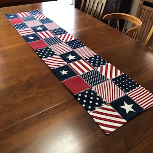 May include: A rectangular table runner with a patchwork design featuring red, white, and blue patterns. The runner includes squares with stars, stripes, and solid colors, evoking an American flag theme. The runner is placed on a wooden table.