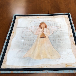 May include: A white angel with gold wings and a trumpet plays a Christmas carol. The angel is on a background of sheet music with the words "O Come All Ye Faithful".