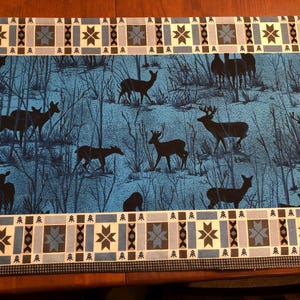 May include: A rectangular placemat featuring a winter scene with deer silhouettes against a blue background. The border has a geometric pattern in shades of blue, gray, and white. The placemat is designed for dining.