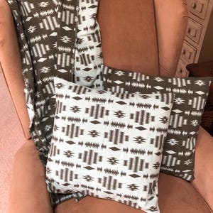 May include: A brown and white patterned throw blanket and two matching throw pillows rest on a peach-colored armchair. The pillows and blanket feature a repeating geometric design. The blanket is draped over the chair and pillows.