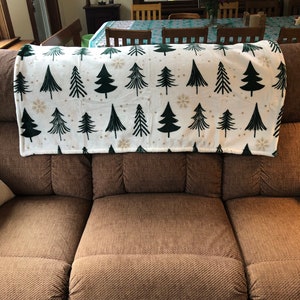 May include: A brown couch with a white throw blanket with a green Christmas tree pattern. The blanket has gold snowflakes and dots.