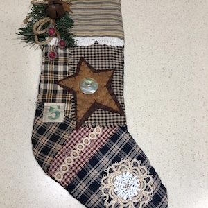 May include: A patchwork Christmas stocking with a star and snowflake design. The stocking features a variety of fabrics in brown, blue, and red plaid patterns. It is decorated with a star, a bell, and faux greenery. A number 5 is visible on the stocking.