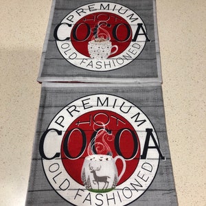 May include: Two gray and white fabric coasters with a red circle in the center. The circle has the text "PREMIUM HOT COCOA OLD FASHIONED" in white lettering. The top coaster has a white mug with a brown drink inside. The bottom coaster has a white mug with a brown drink inside and a deer in the background.