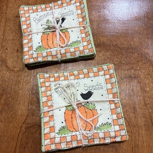 Könnte beinhalten: Zwei quadratische Stoffuntersetzer, die mit Bindfaden zusammengebunden sind, mit einer zentralen Abbildung eines orangefarbenen Kürbisses, eines schwarzen Vogels und dem Text Pumpkin Seeds. Der Rand hat ein orange-weißes Karomuster.