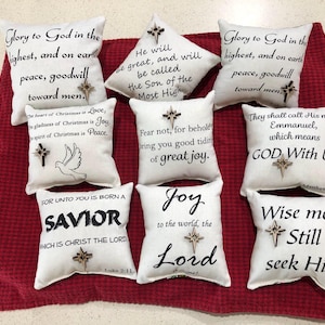 May include: A collection of eight small white square fabric pillows, each featuring black printed religious text and a small wooden star ornament attached to the center. The pillows display various Christmas-themed verses and phrases in different fonts.