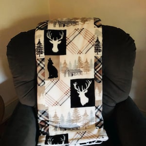 May include: A cozy throw blanket featuring a patchwork design with black and white deer silhouettes, a wolf, and cabin scenes. The blanket has a plaid pattern in shades of brown, black, and white, perfect for a rustic cabin theme.